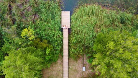 Fliegen-über-Ein-Paar,-Das-Auf-Einer-Holzplattform-Vor-Einem-See-Mit-Klarem-Wasser-Sitzt,-Zwischen-Den-Bäumen,-In-Banyoles,-Katalonien,-Spanien