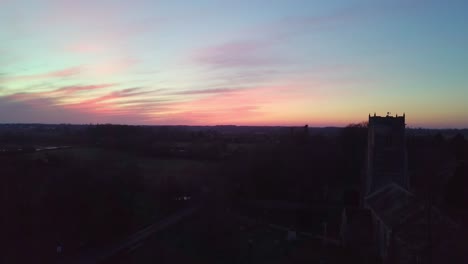 Wunderschöne-Drohnenaufnahme-Bei-Sonnenuntergang,-Die-An-Der-Englischen-Kirche-In-Einem-Ländlichen-Dorf-Vorbeiführt,-Während-Ein-Auto-Die-Straße-Hinunterfährt