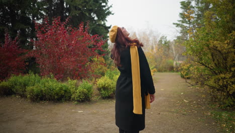 elegant woman in black coat with yellow beret and scarf stands in autumn park, looking around and turning while touching her red hair, vibrant fall colors surround her in a scenic outdoor setting