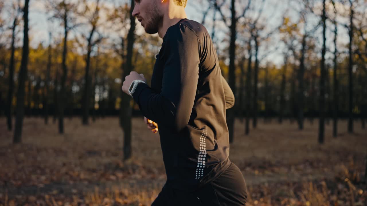 A male athlete in a black sports uniform with curly hair looks at his watch and then continues his run while jogging in the morning in the autumn forest Sunny