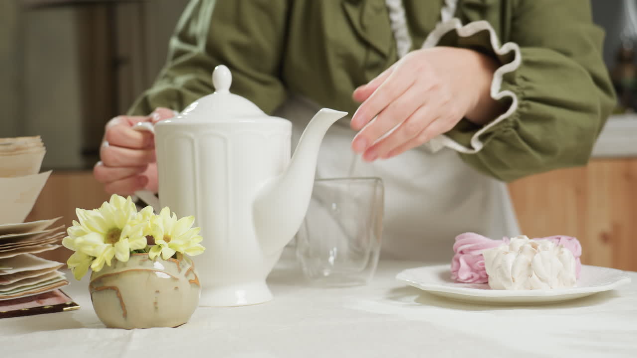 Close up of cozy breakfast table with old book, fresh yellow flowers, dessert plate, and glass mug as female cook places ceramic jug gently before sitting, resting hand under jaw