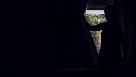 woman in dark bedroom opens the curtains and looks outside