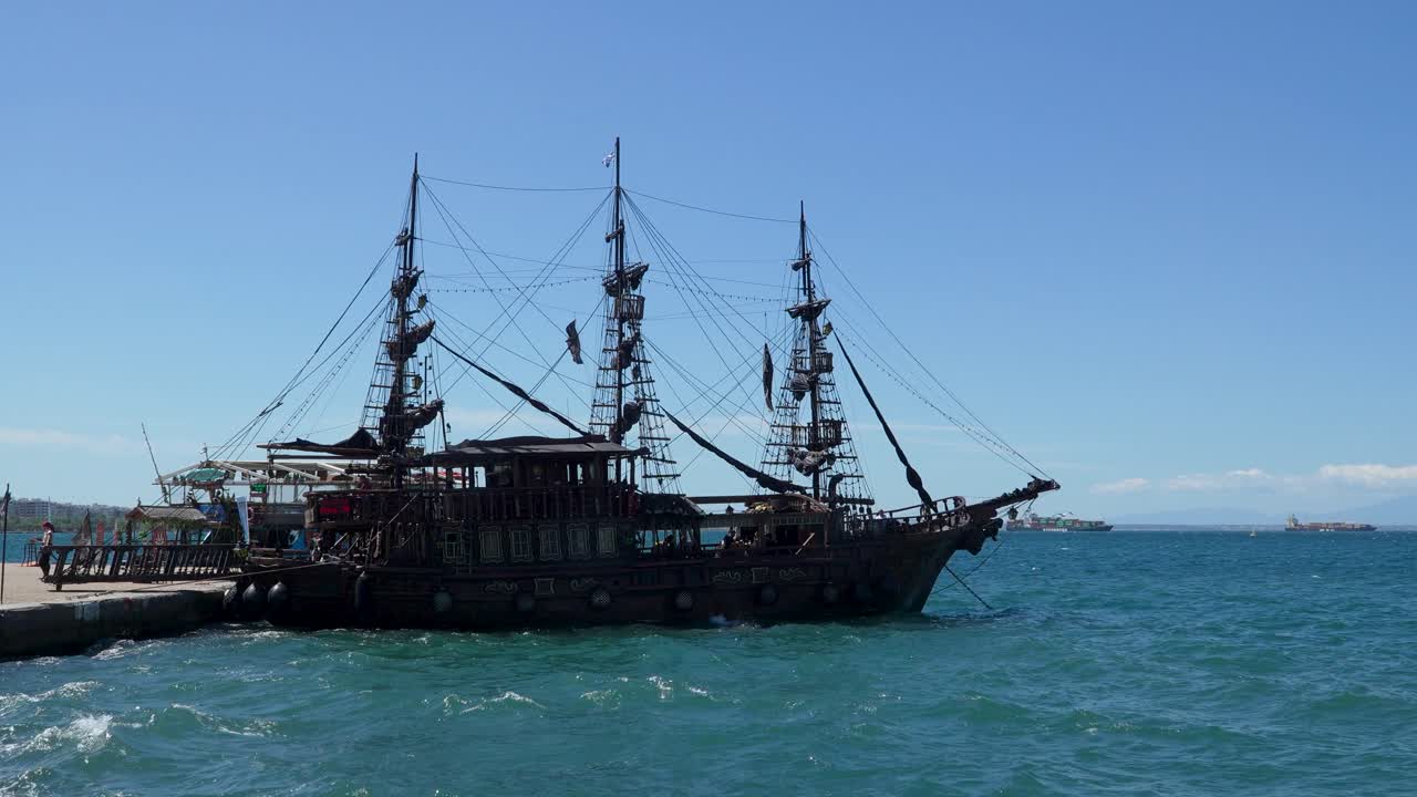 A classic pirate-themed ship with tall masts and black sails is docked at a seaside pier on a clear day, with deep blue waters and distant cargo ships visible on the horizon under a bright sky