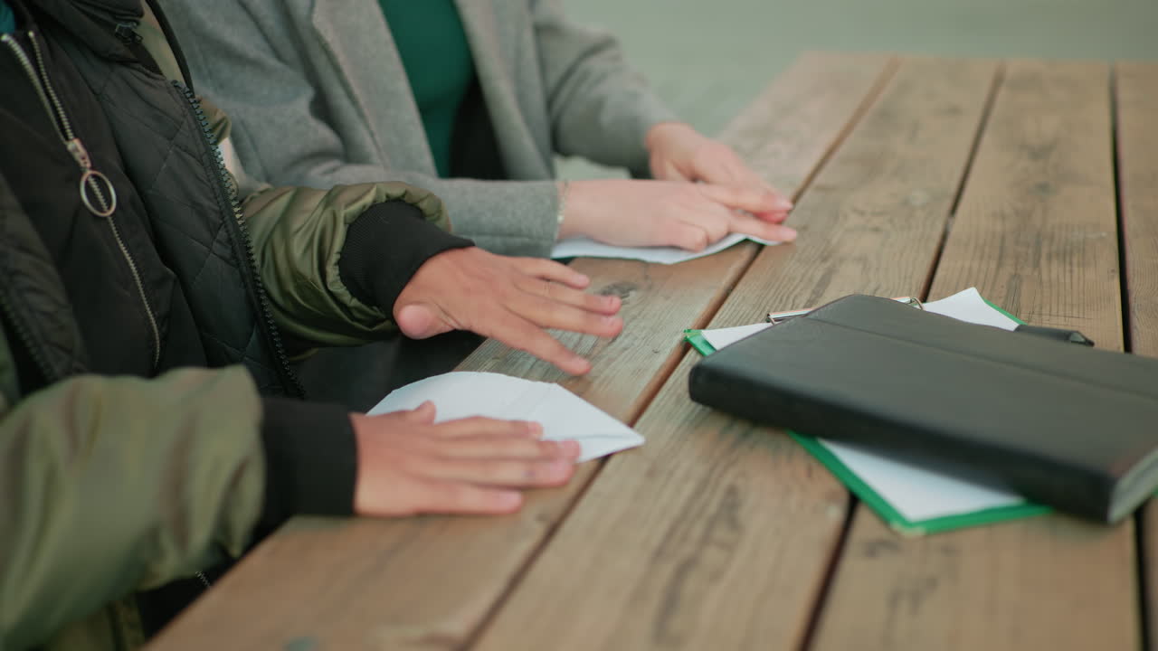 Close up of two people seated outdoors folding paper into kite shape on wooden table, hands carefully working together showing teamwork, learning, and creativity in calm environment