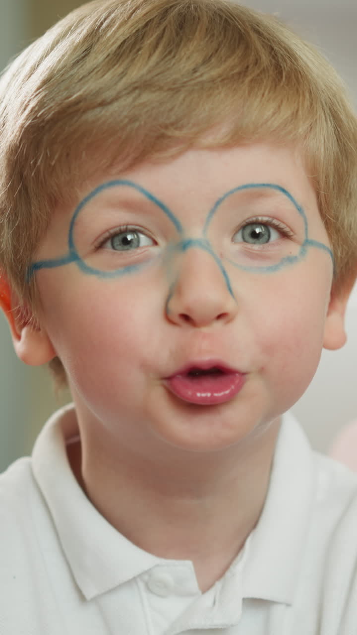 niño preescolar positivo con cara pintada muestra la lengua y aplaude las manos jugando mirando a la cámara. niño travieso se divierte sentado en la habitación en un fondo borroso