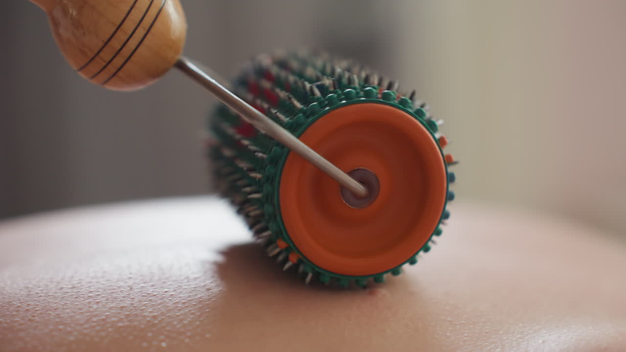 Macro view of colorful massage roller with green bristles, orange rim, and blue tips applying pressure on fair skin back during therapy session, captured with shallow depth and soft blurred background