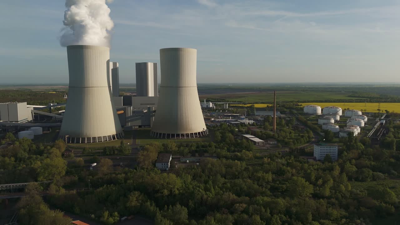 Drone video of Lippendorf coal power station and adjacent rapeseed fields near Leipzig, Germany. Cooling towers, tanks, and bright yellow farmland under clear skies.