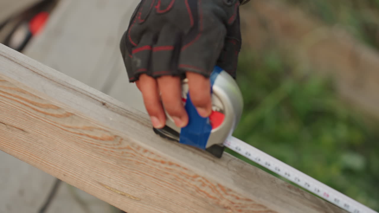 Contractor measuring cut line for repair, gloved hand extends tape over plank and marks precise point for cut, closeup of measurement process, urgent handson fix during outdoor renovation at worksite