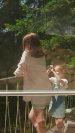 madre posa para una fotografía tomada por su hija con la cámara de pie en el balcón con vistas al denso bosque en un clima soleado de verano. disfrutando de la naturaleza y el apartamento moderno