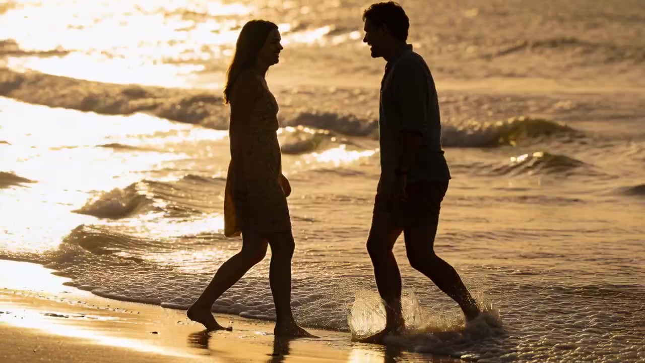 Walking woman in dress approaching man in rolled trousers stepping into surf and meeting on sand