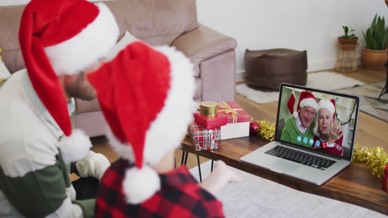 hombre caucásico con hijo usando sombreros de santa en una videochat portátil durante la navidad en casa