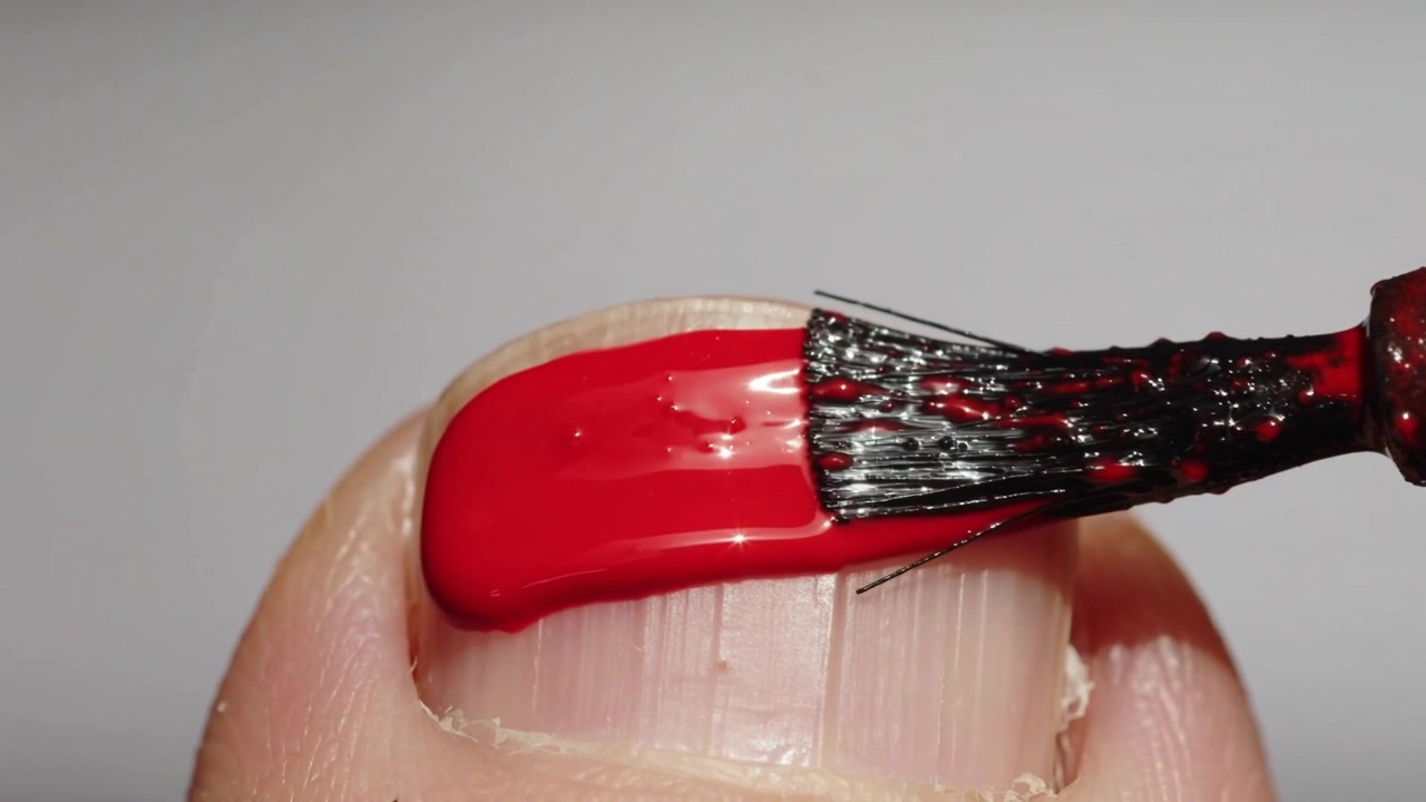 Pincel acercándose y depositando esmalte rojo brillante en la uña del pulgar en primer plano macro, creando una capa suave