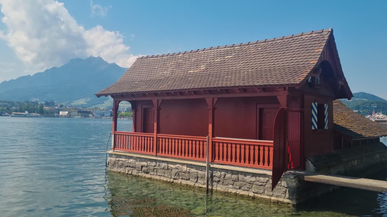 Shot of a small red historic boathouse (Bootshüsli) on Lake Lucerne, captured on a sunny summer day. The calm water reflects the charm of the scene, with majestic Alpine mountains in the background
