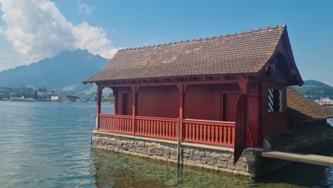 shot of a small red historic boathouse (bootshüsli) on lake lucerne, captured on a sunny summer day. the calm water reflects the charm of the scene, with majestic alpine mountains in the background
