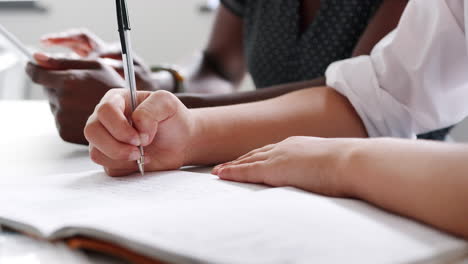 close up of female teacher helping girl high school student with one to one tuition as she writes in book