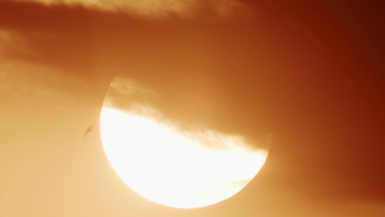 厚い動く雲に部分的に覆われた、鮮やかな黄金色の夕焼け色に輝く太陽のクローズアップ。