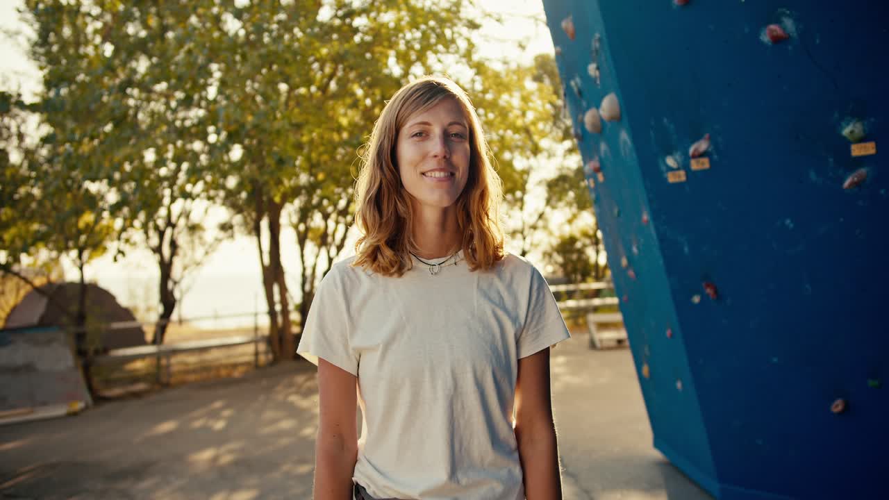 retrato de una feliz chica escaladora de roca rubia en una camiseta blanca posando y mirando a la cámara mientras está cerca de una pared de escalada azul en un día soleado de verano
