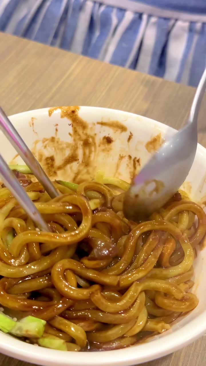 Bacchette e cucchiaio mescolano i noodles jajangmyeon con salsa e verdure in un ambiente da pranzo illuminato calorosamente.