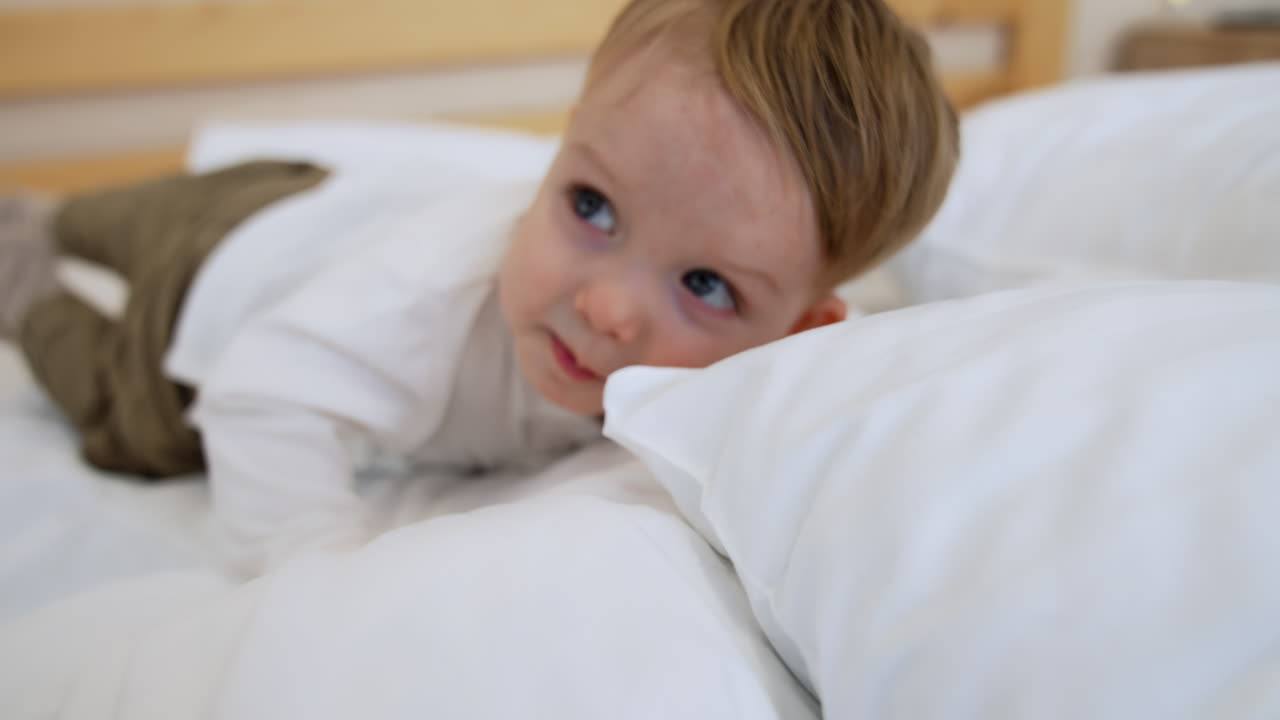 Caucasian toddler kid lies in the pillows on the big bed. Baby boy raises and screams. Close up.
