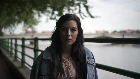 Portrait-of-an-attractive-latina-tourist-with-black-wavy-hair-and-a-jean-jacket-posing-in-a-park-in-London-with-a-view-of-Putney-bridge-behind-her