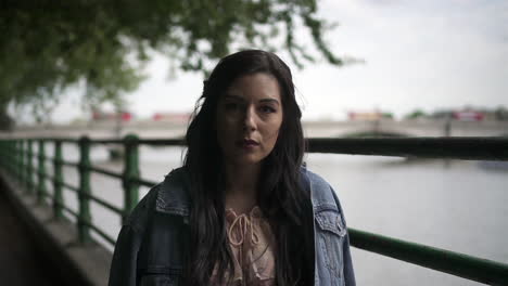 Retrato-De-Una-Atractiva-Turista-Latina-Con-Cabello-Negro-Ondulado-Y-Una-Chaqueta-De-Jean-Posando-En-Un-Parque-En-Londres-Con-Vista-Al-Puente-Putney-Detrás-De-Ella