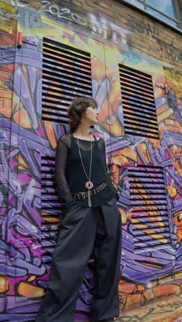 retrato vertical de moda al aire libre de una mujer joven de estilo alternativo inclinándose hacia la cámara contra los muros cubiertos de graffiti de la calle de la ciudad de londres, reino unido, en tiempo real.
