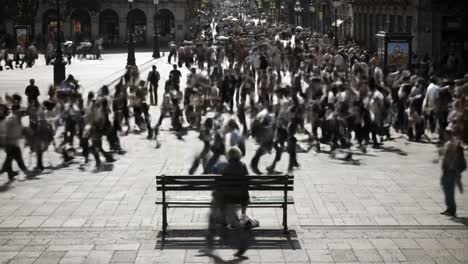 two individuals sit on a bench while a bustling crowd flows past in a vibrant urban square. the atmosphere is energetic, showcasing daily life and human interaction in the city.