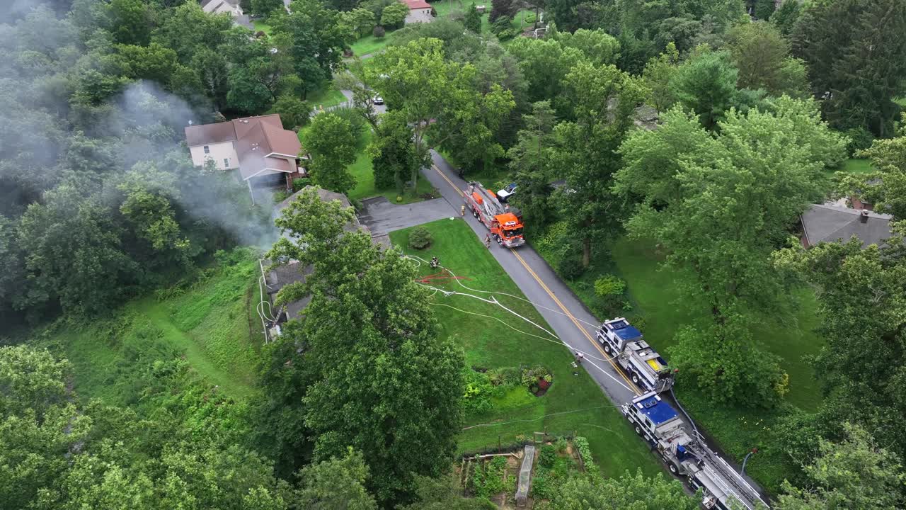 Several fire trucks extinguishing burning house with rising smoke. Aerial top shot. Green suburb neighborhood in summer
