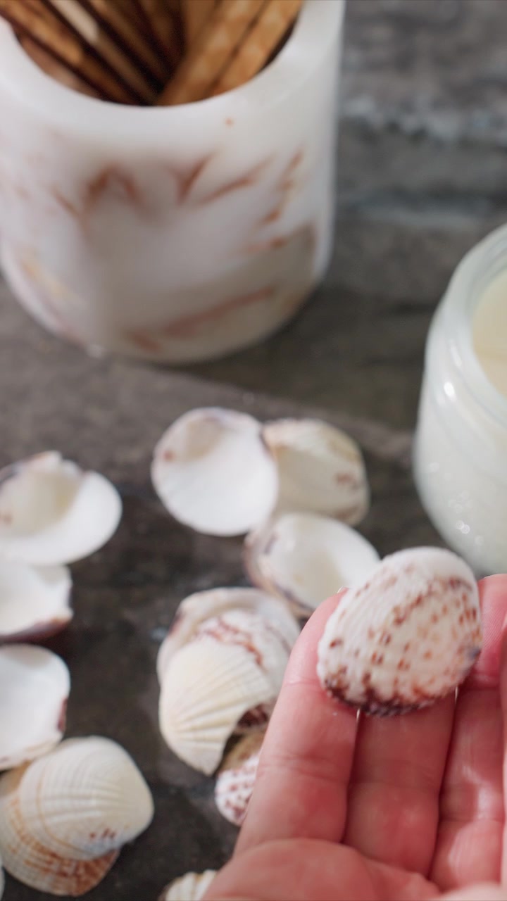 Close up of hand gently picking and examining small seashells on dark stone surface. shells display white with brown patterns while background includes blurred candles in glass jars. Soft