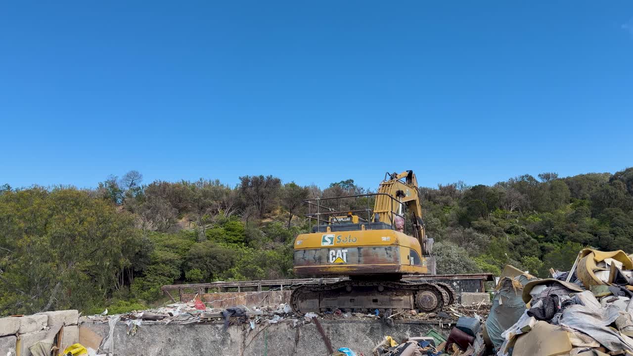 Excavator operates at landfill, moving rubbish among trees and hills under clear blue sky