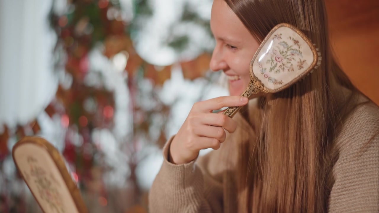 Donna che si acconcia i capelli. Ambiente interno casual con donna sorridente. Donna gioiosa che si spazzola i capelli lunghi al chiuso. Donna rilassata che si acconcia con entusiasmo i suoi lunghi capelli vicino a uno specchio decorativo al chiuso