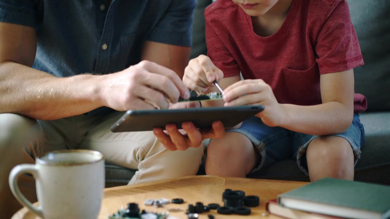 Leaning adult male guiding child using screwdriver on couch to assemble circuit board, with tablet