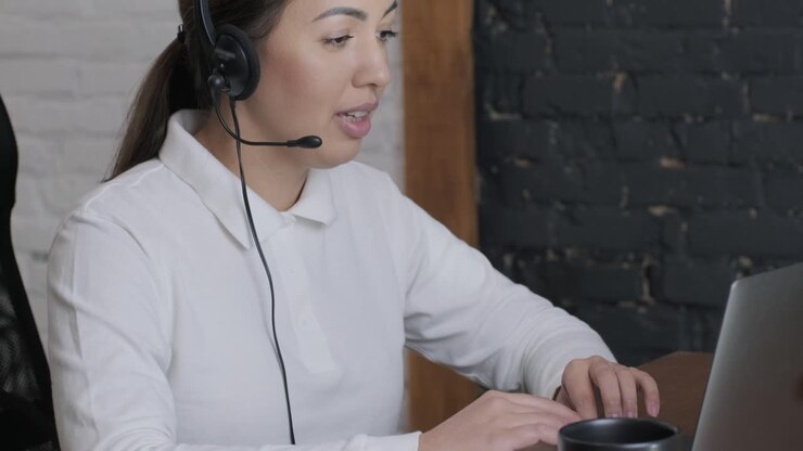 Smiling woman call centre operator, customer support agent wears headset consults client online typing on laptop, talk in internet computer chat, helpline operator secretary make conference video call