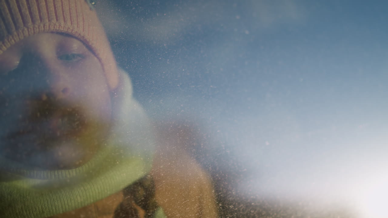 핑크색 뜨개질 모자와 스카프를 입은 어린 소녀가 안개 차 창문에 글을 쓰고, 얼굴을 유리창에 렀습니다.