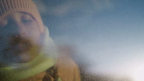 a young girl in a pink knit hat and scarf writing on a foggy car window, with her face pressed against the glass