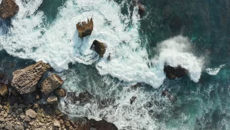 aerial top down view of ocean waves crashing into rocky shore with large rocks