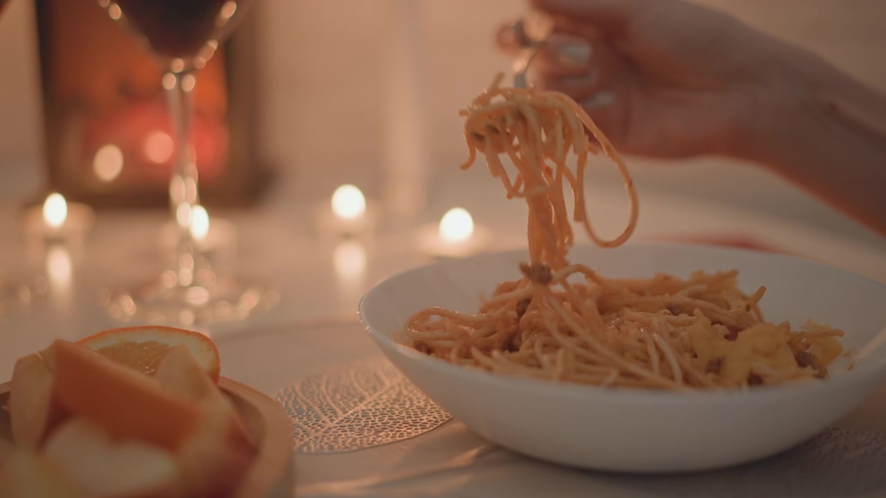 Primo piano di una mano che si protende verso una forchetta per avvolgere gli spaghetti in una ciotola di ceramica bianca, catturando il movimento attivo del mangiare e i fili di pasta sospesi contro uno sfondo caldo a lume di candela con pane e vino nelle vicinanze.
