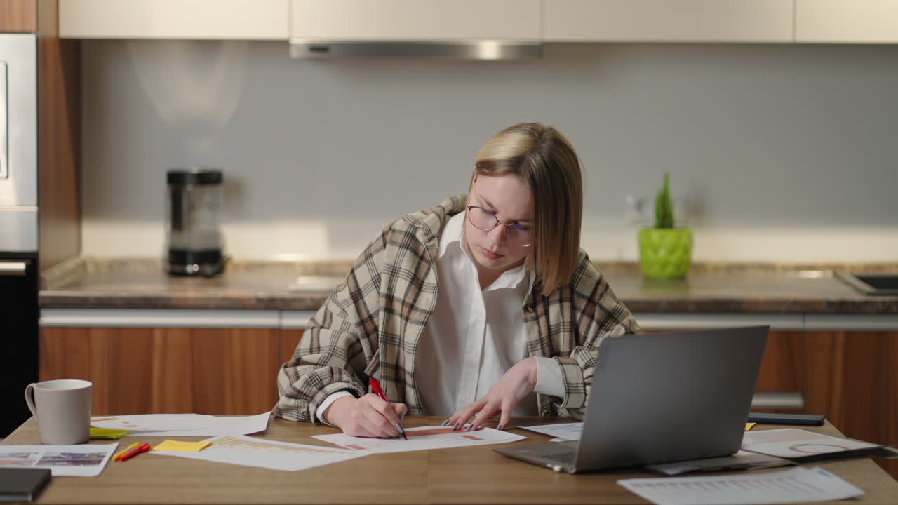 una mujer con gafas trabaja remotamente desde casa sentada en una mesa con una computadora portátil y un bolígrafo marca los datos en el gráfico. estudiante de aprendizaje a distancia en casa para realizar una tarea en economía