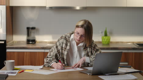 Una-Mujer-Con-Gafas-Trabaja-Remotamente-Desde-Casa-Sentada-En-Una-Mesa-Con-Una-Computadora-Portátil-Y-Un-Rotulador-Marca-Los-Datos-En-El-Gráfico.-Estudiante-A-Distancia-En-Casa-Para-Realizar-Una-Tarea-De-Economía