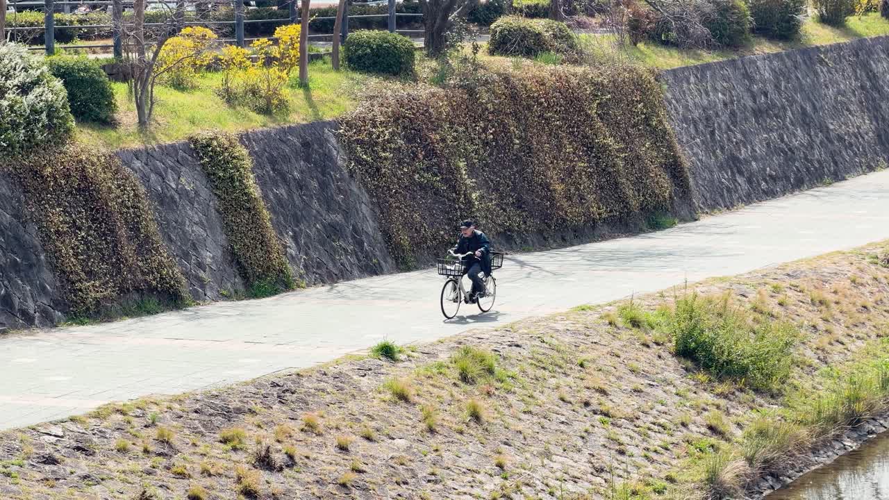 ワイドショット：舗装された川岸の小道で自転車に乗る高齢の男性を捉える。