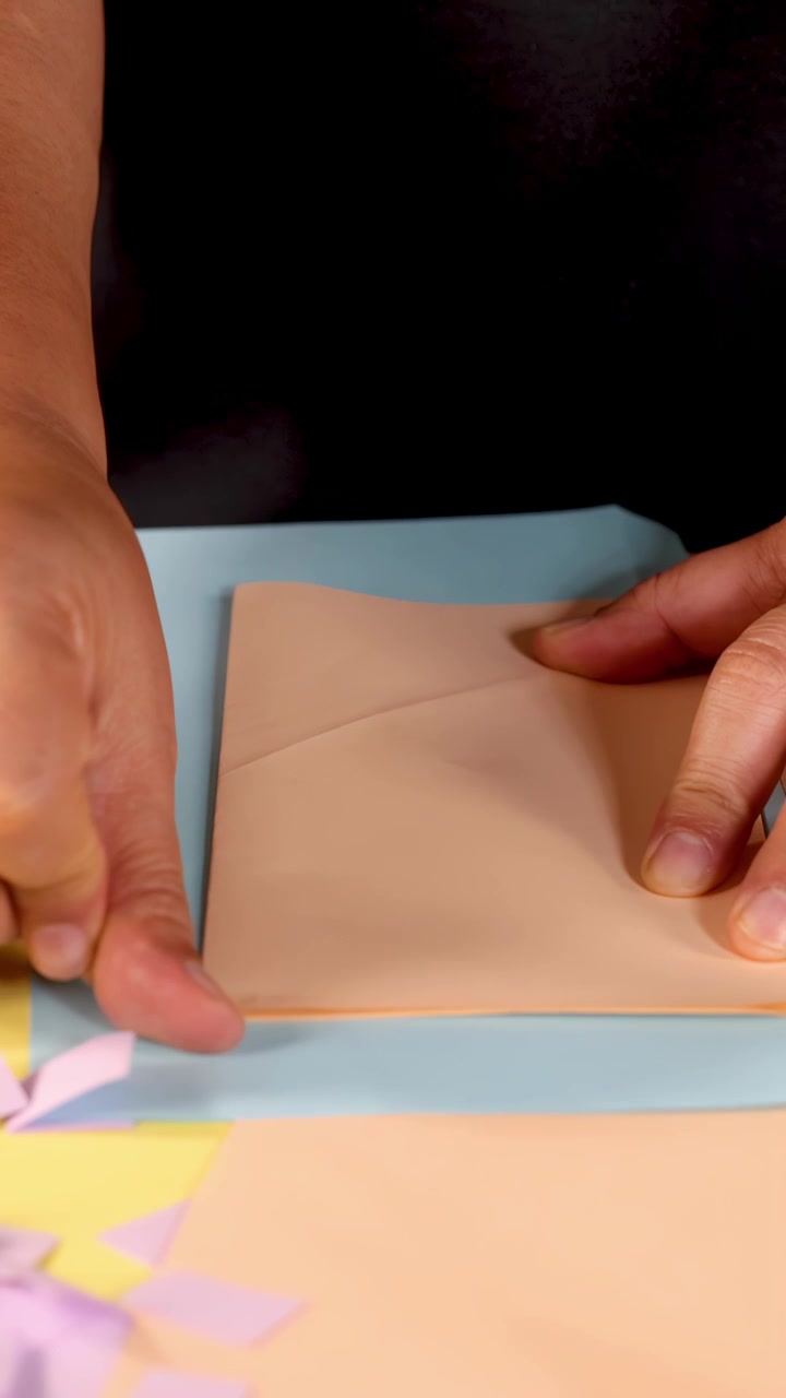 Voksne hænder folder forsigtigt pastel origami-papir, nærbillede, under blød studiebelysning, statisk kamera.