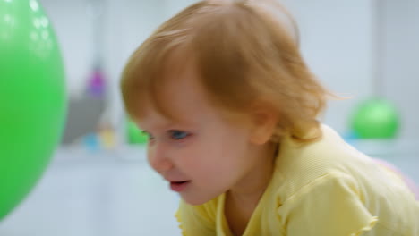 toddler in yellow top lying down, turning to look up with a curious expression beside a large green exercise ball, vivid colors and innocent gaze captured in a gym setting