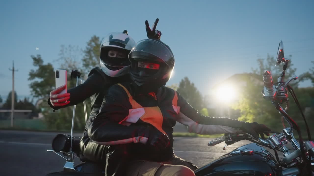 Lady in white helmet takes selfie with husband on motorcycle at sunset, husband in black helmet poses while she makes peace sign gesture, evening road and trees create scenic background
