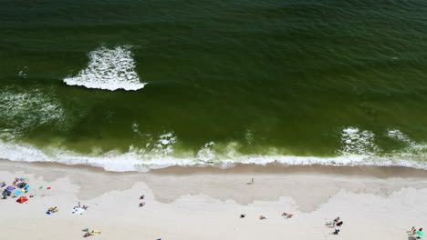 tilt-down-bird's-eye-view-of-the-shoreline-as-the-ocean-waves-crash-against-the-shore-and-people-walking-by-and-enjoying-the-day-and-sun,-while-the-drone-camera-hovers-in-one-place