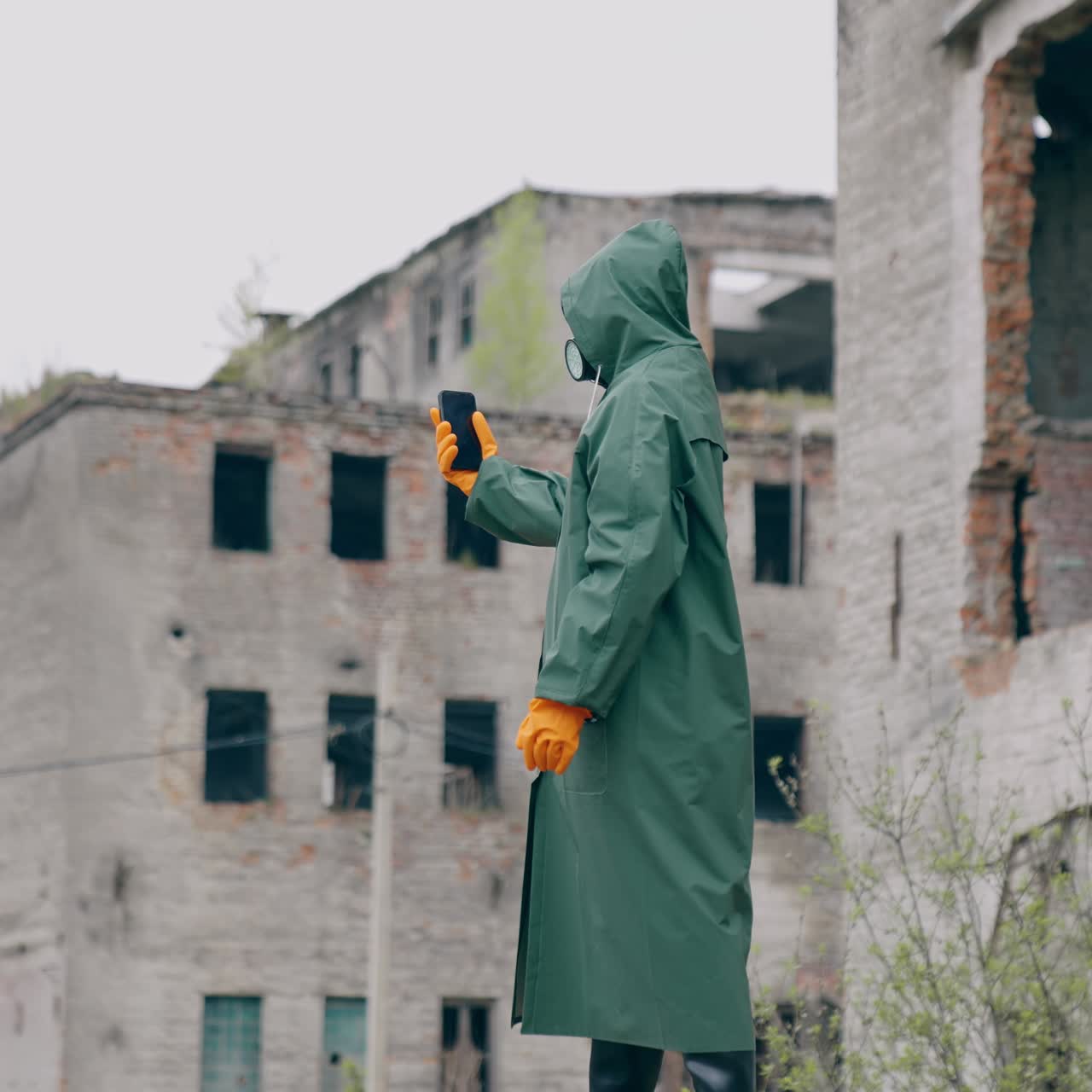 Human in protective suit and gas mask in abandoned place. Survivor holding a mobile phone while standing in ruined city. Post-apocalyptic world after a nuclear war