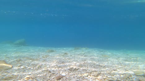 underwater footage of a seabed with white pebbles, blue sea and reflections on the surface