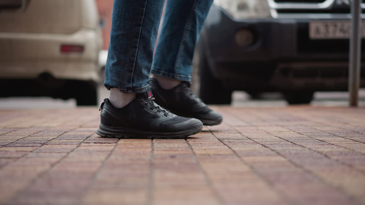 Side view of man wearing black winter coat and blue jeans walking along tiled urban walkway beside parked cars and brick buildings with focus on sneaker motion and footstep pattern during cold day