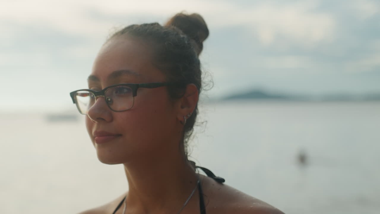 Closeup woman smiling by ocean sunlit threequarter portrait of confident young woman with glasses and topknot, warm golden light, subtle smile, seaside islands on horizon, lifestyle travel vibe, model