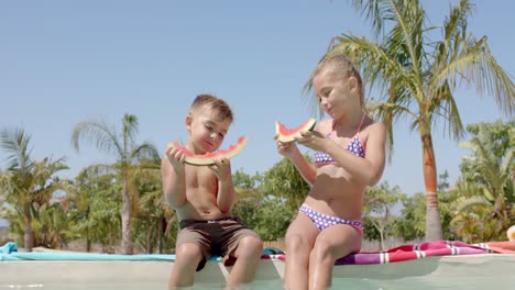 happy caucasian siblings eating watermelon at swimming pool at beach house