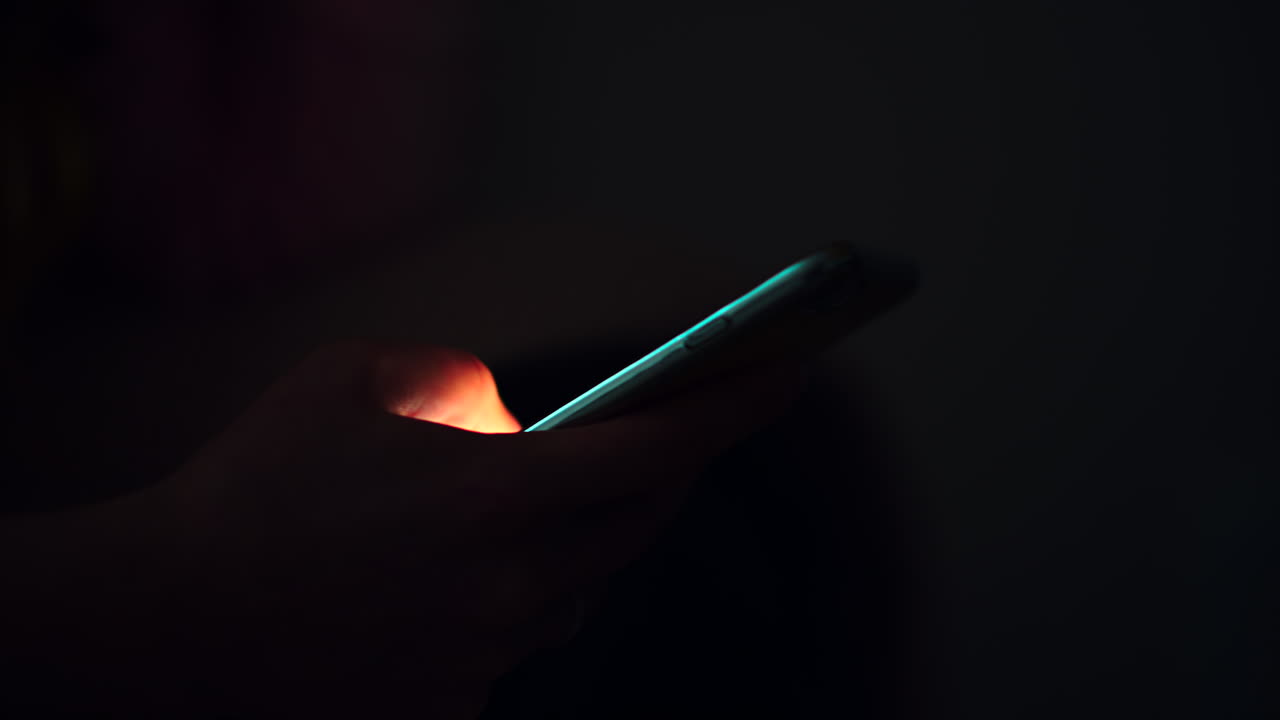 A person is holding a smartphone while sitting in a dark space, glowing screen illuminating their hand as they browse messages in the late evening
